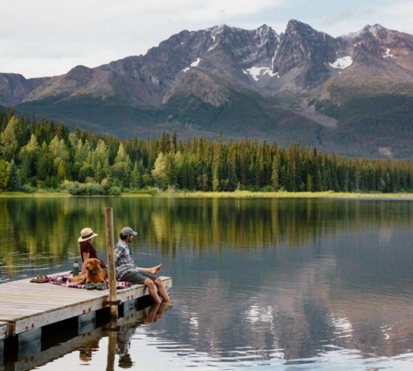 Zwei Personen sitzen auf einem Steg am See mit einem Hund, umgeben von Bergen und Bäumen. Kanada-Rundreise