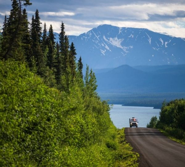 Eine Straße entlang des Dease Lake mit Bergen im Hintergrund und Bäumen auf beiden Seiten. Kanada-Rundreise