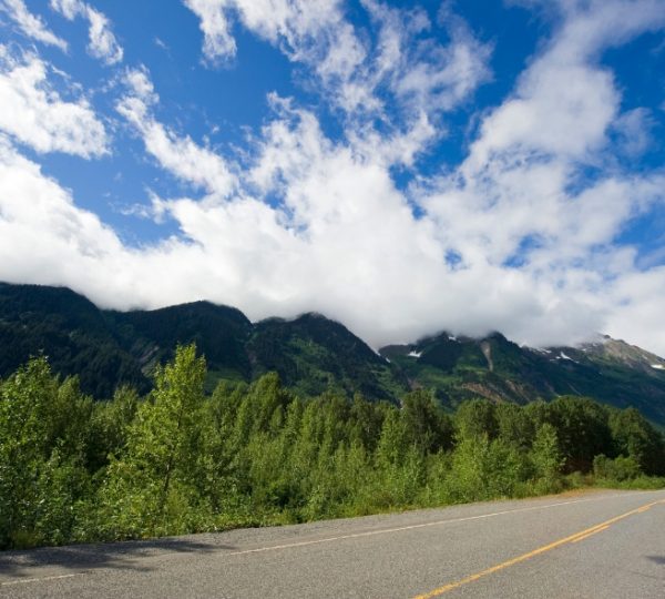 Eine Straße in Westkanada mit bewaldeten Bergen und wolkigem Himmel im Hintergrund. Kanada-Rundreise