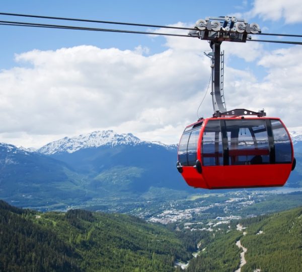 Rote Gondelbahn schwebt über einer grünen Berglandschaft mit schneebedeckten Gipfeln im Hintergrund. Kanada-Rundreise
