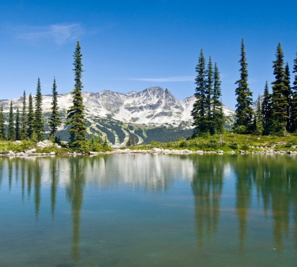 Berglandschaft mit einem klaren See und hohen Nadelbäumen im Vordergrund. Kanada-Rundreise