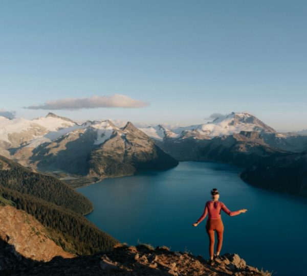 Person steht auf einem Berg mit Blick auf einen See und schneebedeckte Berge in Westkanada. Kanada-Rundreise