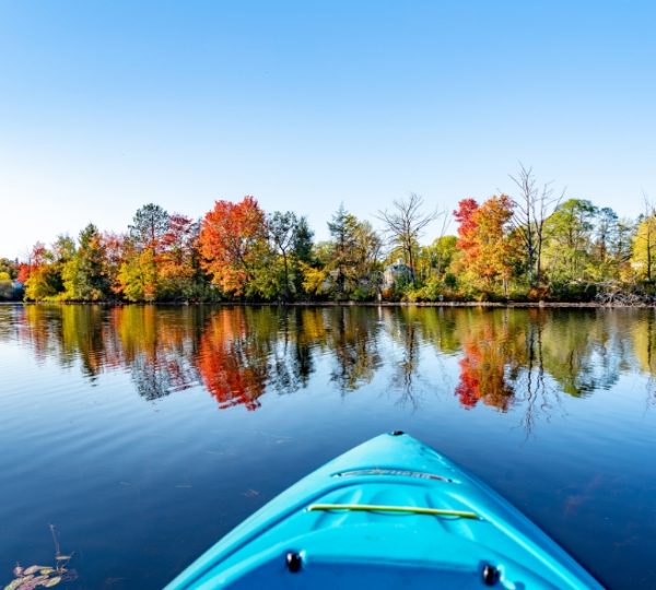 Blaues Kajak im Vordergrund auf einem ruhigen Gewässer, umgeben von bunten herbstlichen Bäumen und klarem Himmel. Kanada-Rundreise