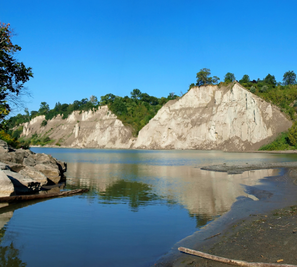 Klippen mit grüner Vegetation am Ufer eines ruhigen Gewässers unter klarem Himmel. Kanada-Rundreise