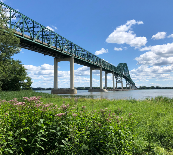 Grüne Brücke mit Stahlkonstruktion, die über einen Fluss führt, umgeben von Pflanzen und Wasser. Kanada-Rundreise