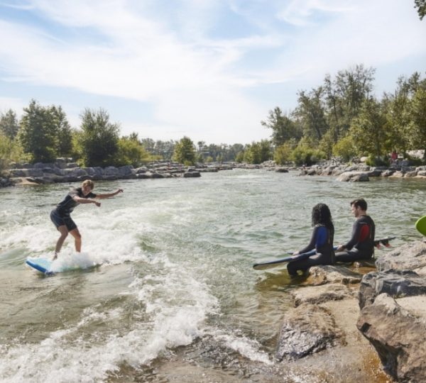 Surfer, der auf einer Welle im Harvie Passage in Calgary surft, während andere im Wasser sitzen. Kanada-Rundreise