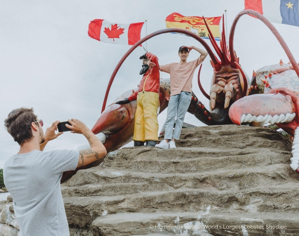 Zwei Personen posieren vor dem größten Hummer der Welt in Shediac, umgeben von verschiedenen Flaggen, während eine dritte Person ein Foto macht. New Brunswick