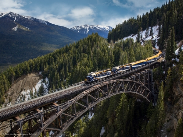 Rocky Mountaineer Zug, der über eine Brücke in den Rocky Mountains fährt, umgeben von Bäumen und Bergen. Kanada Zugreise