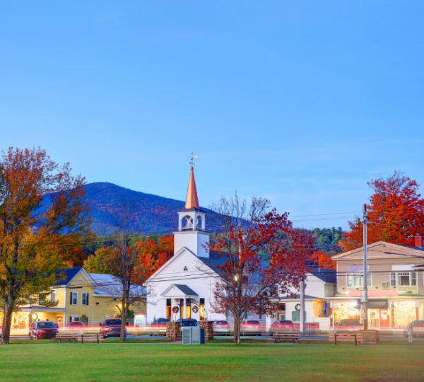 Eine herbstliche Kleinstadtansicht in North Conway mit einer weißen Kirchturmkirche im Zentrum, umgeben von rotem und orangefarbtem Laub, vor Bergen unter klarem Himmel Kanada-Rundreise