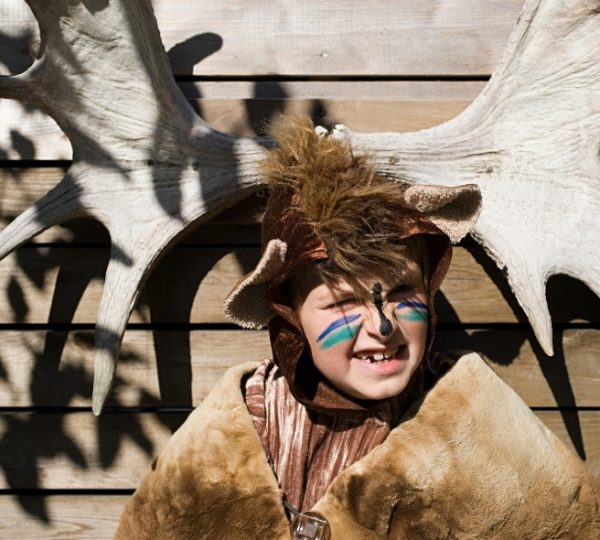 Ein Kind trägt ein braunes Moose/Moose-Kostüm mit Geweih-Aufsetzern und bunten Gesichtsblicken vor einer Holzwand; Shadow-Entwürfe der Geweih-Silhouette im Hintergrund Kanada-Rundreise