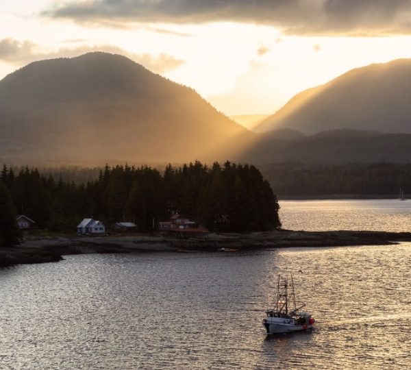 Sunset über einer Küstenlandschaft mit Boot im Vordergrund, kleine Häuser am Ufer, dichte Waldgürtel und Bergkette im Hintergrund – Alaska-Kreuzfahrt mit NCL Encore Kanada-Rundreise
