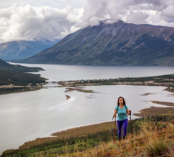Wanderin mit Trekkingstöcken steht auf grünem Hang über einem großen See, bergige Landschaft im Hintergrund, Wolken am Himmel Kanada-Rundreise