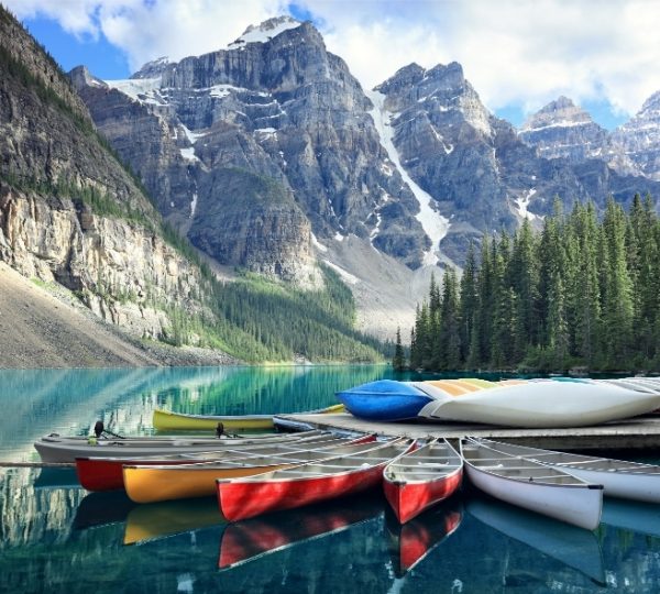 Blaue Moraine Lake in Banff, Kanada, mit bunten Kanus am Ufer, umgeben von dichtem Wald und majestätischen, schneebedeckten Berggipfeln Kanada-Rundreise