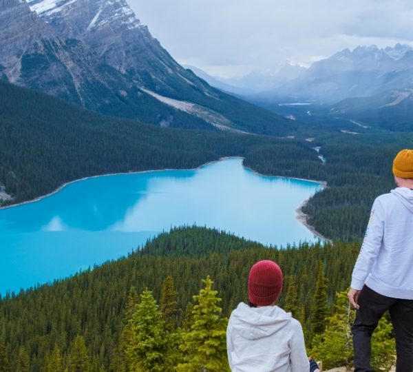 Blauer Peyto Lake in Banff-Nationalpark umgeben von Wald und Gipfeln; zwei Personen stehen am Aussichtspunkt und blicken auf den See Kanada-Rundreise