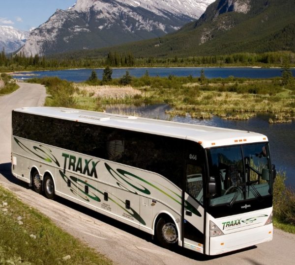 Weißer TRAXX Reisebus fährt auf einer unbefestigten Straße nahe einem See, umgeben von Wald und Bergen; Bus mit grünen Designelementen. Kanada-Rundreise