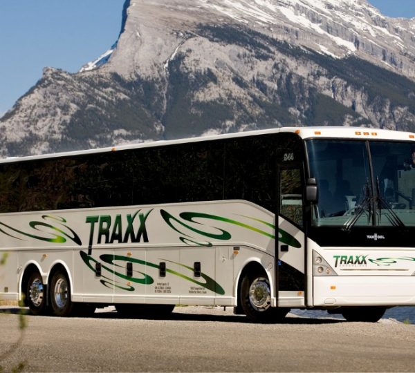 Weißer TRAXX-Reisebus mit grünen Schwunglinien parkt am Ufer eines Sees, vor imposanten Bergketten und Wald im Hintergrund, klare, sonnige Szenerie Kanada-Rundreise