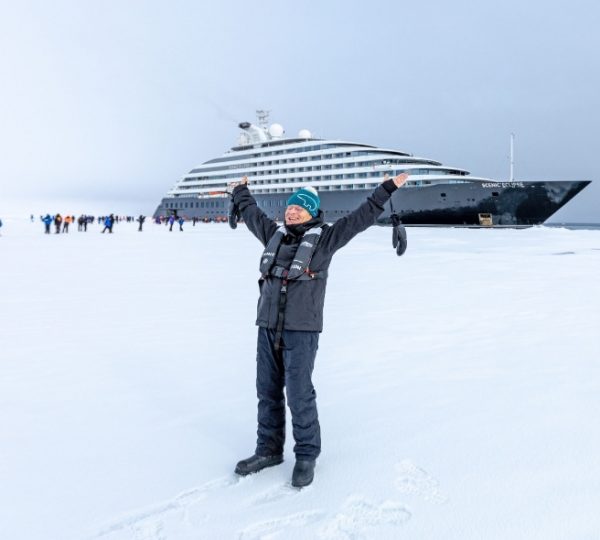 Eine Person in Winterkleidung jubelt vor dem majestätischen Kreuzfahrtschiff Scenic Eclipse, das auf einer eisbedeckten Küste der Hudson Bay vor Ankunft verweilt, umgeben von weiter, verschneiter Landschaft. Kanada-Rundreise