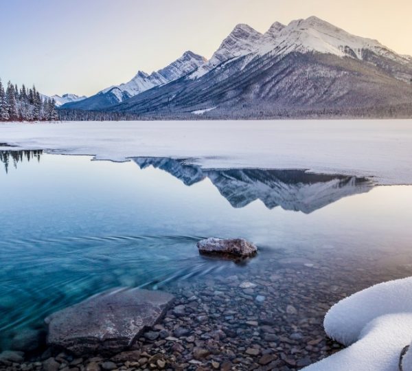 Kananaskis Lake im Winter mit verschneiten Bergen und Wald am Ufer; das ruhige Wasser spiegelt die Berglandschaft, leichter Sonnenaufgang am Horizont Kanada-Rundreise