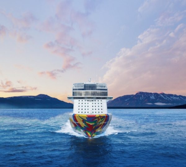 Alaska Kreuzfahrtschiff NCL Encore fährt durch ruhige Meereswellen vor einer Bergkette bei Sonnenuntergang; farbige Schiffsnase im Vordergrund Kanada-Rundreise