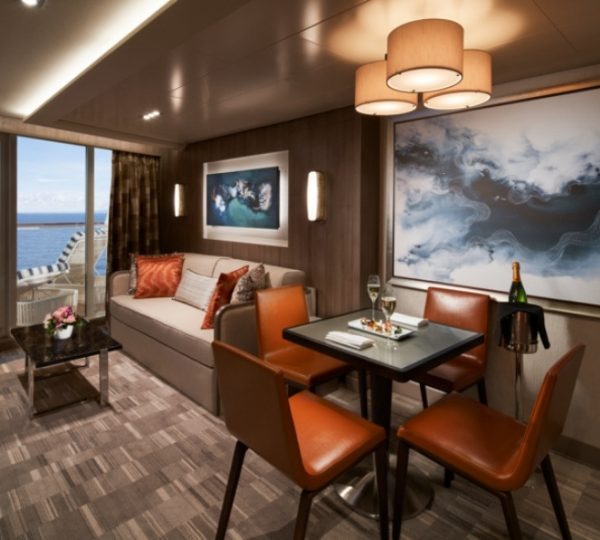 Innenaufnahme einer Alaska-Kreuzfahrt-Kabine mit Meerblick: braune Holzwandverkleidung, orange Lederstühle, Sofa, Glastisch, Balkonblick aufs Meer und dekorative Bilder Kanada-Rundreise