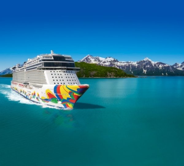 Bunt bemaltes Kreuzfahrtschiff der Norwegian Cruise Line (NCL) segelt durch türkisblaues Wasser vor einer schneeigen Berglandschaft in Alaska. Kanada-Rundreise