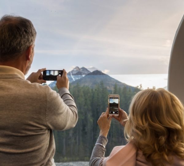 Zwei Reisende im Zugabteil fotografieren eine Berglandschaft durch Fenster; im Hintergrund hohe Berge, Wald und ein offener Himmel Kanada-Rundreise