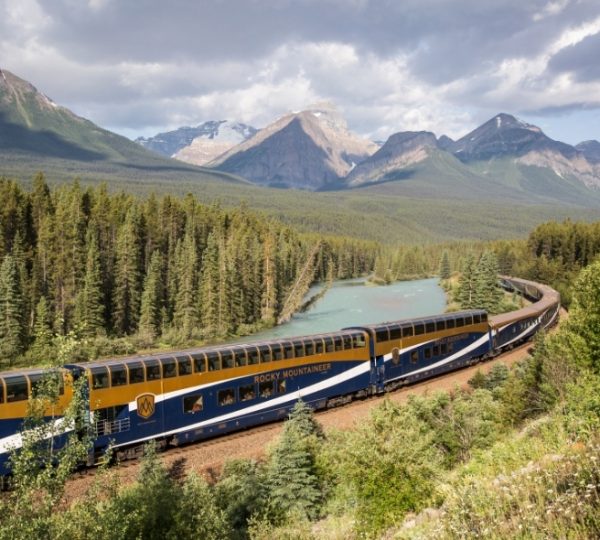 Der Rocky Mountaineer Zug in blau-gelb zieht durch eine Berglandschaft Kanadas, begleitet von einem türkisfarbenen Fluss und grünen Wäldern, majestätische Berge im Hintergrund Kanada-Rundreise