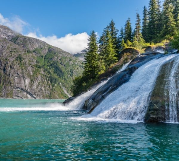 Rocky Mountaineer Landschaft mit türkisfarbenem Wasser, Wasserfall an felsiger Küste, umgeben von grünem Wald und imposanten Bergen – Vancouver und Alaska Kreuzfahrt Kontext Kanada-Rundreise