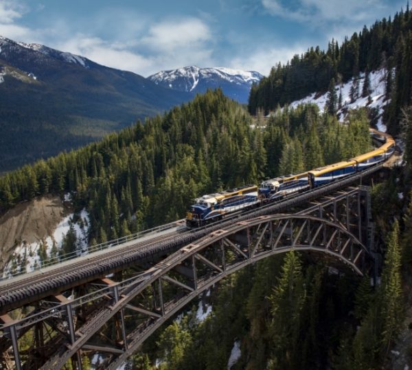 Rocky Mountaineer Zug in gelb-blauer Färbung über eine hohe Stahlbrücke durch bewaldete Berglandschaft mit Schneeresten – Vancouver und Alaska Kreuzfahrt Hinweis Kanada-Rundreise