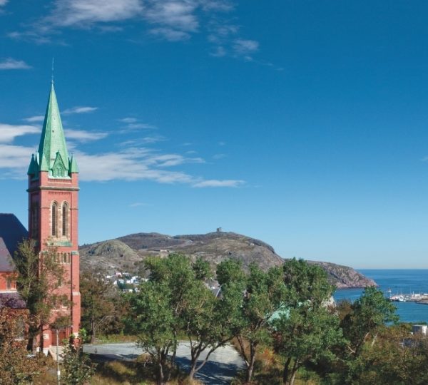 Saint John roter Backstein-Kirchturm mit grüner Turmspitze vor Küstenlandschaft, Bäume im Vordergrund, blauer Himmel und Meer im Hintergrund Kanada-Rundreise