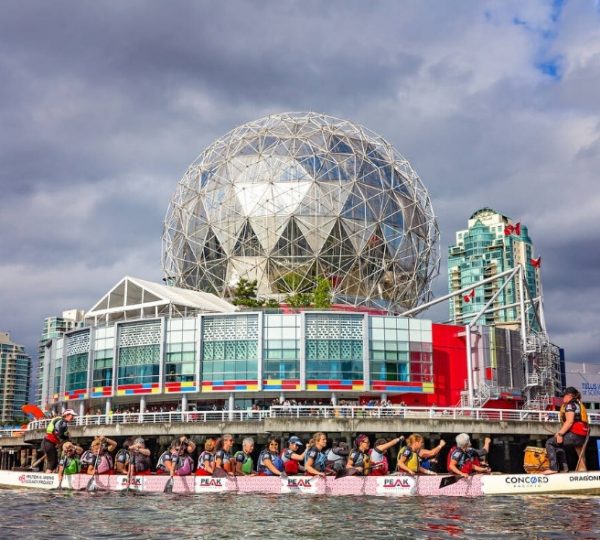 Boote mit Paddlern im Vordergrund vor der silbern schimmernden geodätischen Kugelstruktur des Science World in Vancouver, Kanada, bei wolkigem Himmel und ruhigem Wasser Kanada-Rundreise