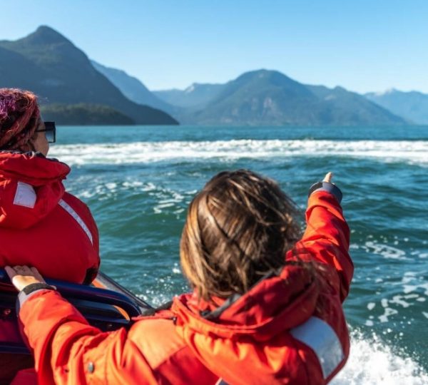 Vancouver-Wal-Beobachtung: Zwei Personen in roten Jacken stehen auf einem Boot und sehen aufs Meer, Berge im Hintergrund, Frühlingssonne, Wellen und Blick aufs Wasser Kanada-Rundreise