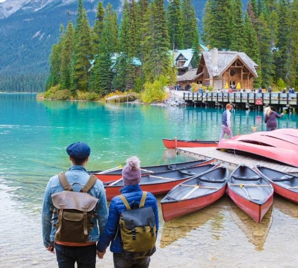 Blaugrünes Emerald Lake Wasserfront mit roten Booten am Ufer, umliegender Wald und eine Holzhütte auf dem Pier; Paare mit Rucksack im Vordergrund Kanada-Rundreise