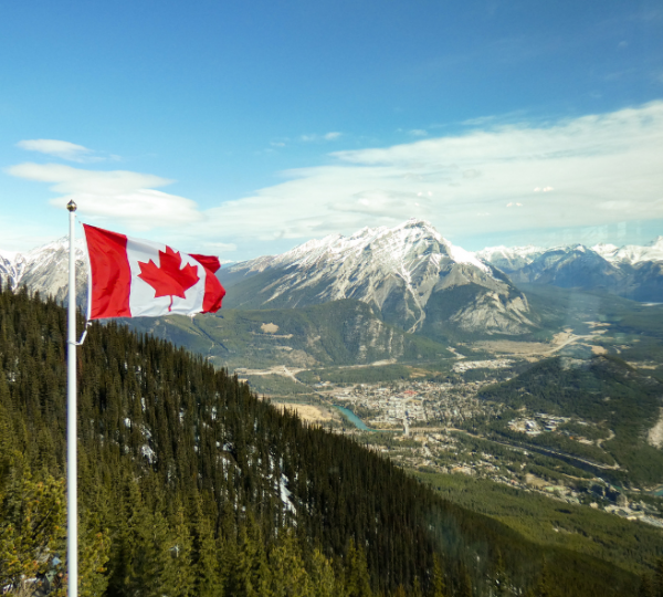 Kanadische Flagge weht auf einem Mast vor einer Berglandschaft mit schneebedeckten Gipfeln, grünen Wälder und einer Stadt im Tal; blauer Himmel Kanada-Rundreise