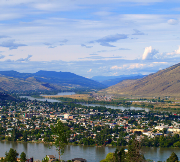 Landschaftsaufnahme in Kamloops, British Columbia: breiter Fluss im Vordergrund, ufernahe Bebauung mit Bäumen, sowie sanfte Berge im Hintergrund unter blauem Himmel Kanada-Rundreise