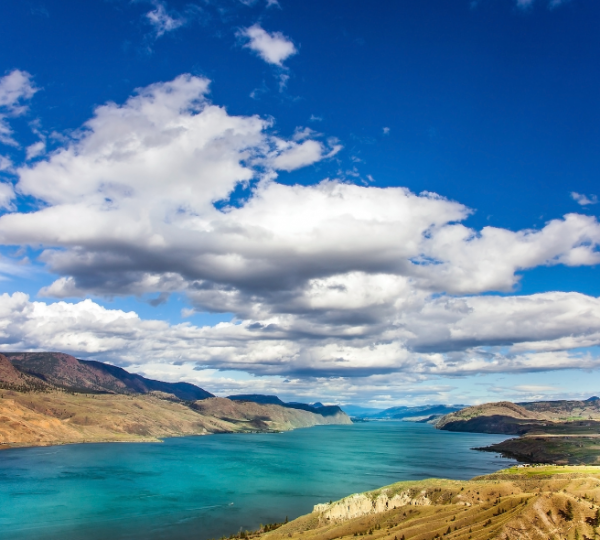 Kamloops Panorama mit blauem Fluss- und Uferlandschaft, grüne Hügeln im Hintergrund, dramatische Wolken am klaren Himmel Kanada-Rundreise