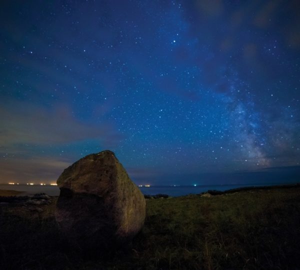 Nachtaufnahme an der Küste von L’Anse-au-Clair mit einem großen Felsen, Sternenhimmel, ruhigem Wasser und grünem Ufer Kanada-Rundreise