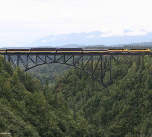 Alaska Railroad – gelber Zug fährt über eine Eisenbahnbrücke, umgeben von dichtem Wald, mit Bergkette im Hintergrund und klarer Ferne. Kanada-Rundreise