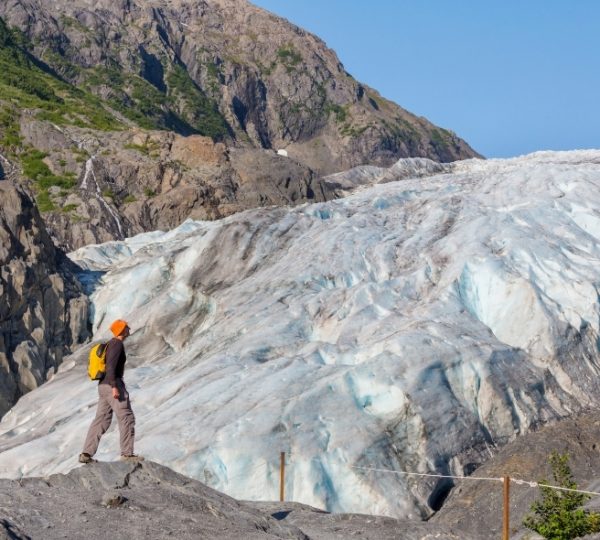 Wanderer mit gelbem Rucksack blickt auf den blauen Hubbard-Gletscher in Alaska; felsige Berglandschaft im Hintergrund, klare Himmel, Eis und Gestein kontrastieren Kanada-Rundreise