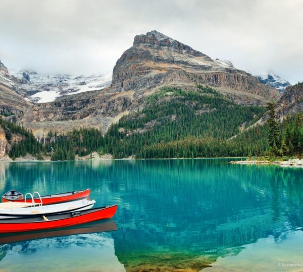 Blaues Wasser eines Bergsee im Banff/Jasper Gebiet, Bootsreihen am Ufer, umgeben von dichtem Nadelwald und hohen Berggipfeln Kanada-Rundreise