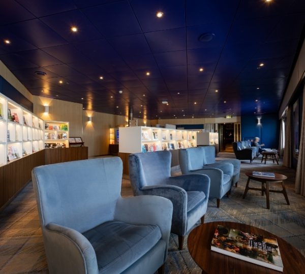 Innenansicht der Zuiderdam-Lounge mit blauen Sesseln, Regalwand mit Magazinen, dunkle Holztische, große Fensterfronten und warme Beleuchtung Kanada-Rundreise
