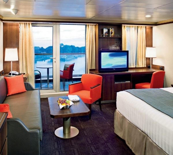 Innenansicht einer Kreuzfahrt-Kabine auf Nieuw Amsterdam mit Balkon, Sofa, TV, Tisch, orangefarbene Stühle und Blick auf ruhiges Wasser; warme Holzoptik und gemütliche Details Kanada-Rundreise