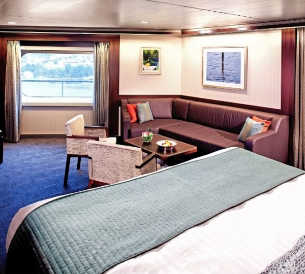 Interior der Kabine auf der Nieuw Amsterdam mit blauem Bettüberwurf, braunen Sofa, bunten Kissen, Fenster mit Blick auf Wasser, gemütlicher Sitzecke Kanada-Rundreise