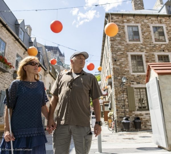 In Québec City führen orange Ballons durch sonnige Altstadtgassen zwischen Natursteinfassaden, während Reisende gelassen an einer historischen Kulisse entlanggehen. Kanada-Rundreise