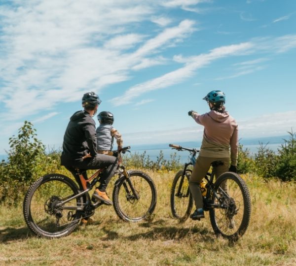 Mountainbiker fahren durch grünes Gras mit weitem Blick in die Ferne, unter blauem Himmel und vor der Landschaft des Le Massif de Charlevoix in Québec. Kanada-Rundreise