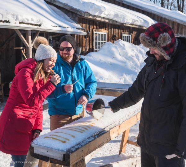 Winterritual in Quebec: Ahornsirup auf Schnee zwischen verschneiten Holzhütten, warm gekleidete Reisende genießen die süße Tradition. Kanada-Rundreise