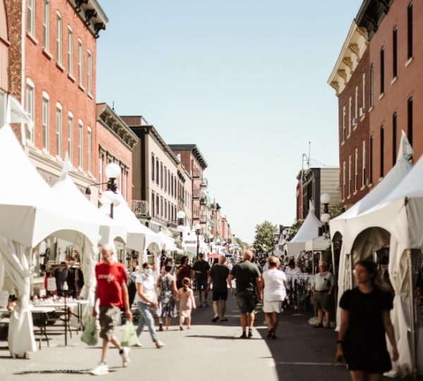 Blick auf eine belebte Einkaufsstraße mit roten Backsteinfassaden und weißen Marktzeltplanen in Saint-Hyacinthe, Quebec, mit vielen Spaziergängern. Kanada-Rundreise