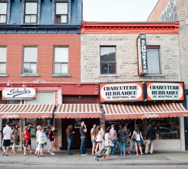 Montréal mit roter Markisenfront und Backsteinfassade, vor den Schaufenstern von Charcuterie Hebraique und Chez Schwartz, belebte Straßenszene. Kanada-Rundreise