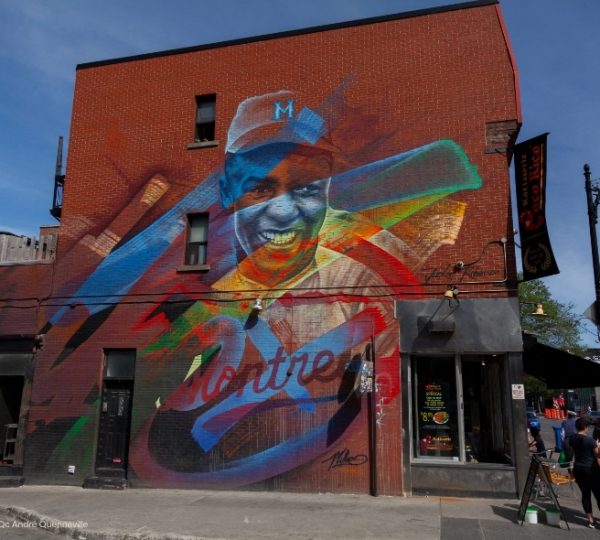 Buntes Mural auf einer Backsteinwand in Montreal mit Gesichtern, kräftigen Farben und dynamischen Formen vor blauem Himmel Kanada-Rundreise