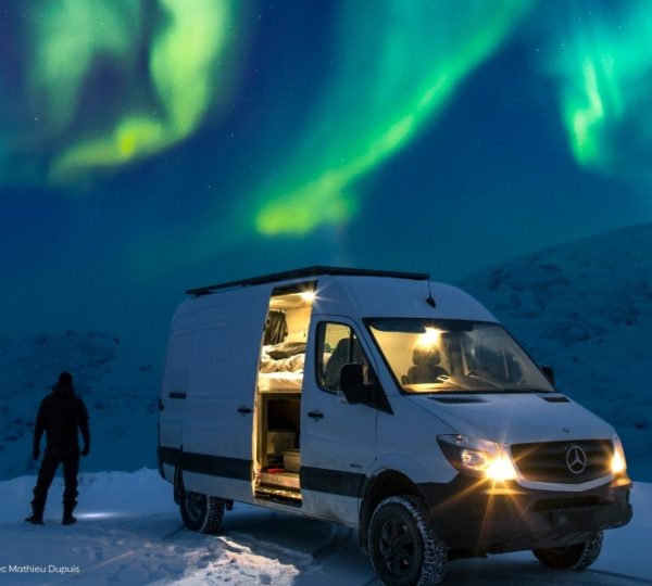 Grüne Nordlichter über schneebedeckter Landschaft in Quebec bei Nacht, im Hintergrund verschneite Hügel und eine beleuchtete Unterkunftsroute. Kanada-Rundreise